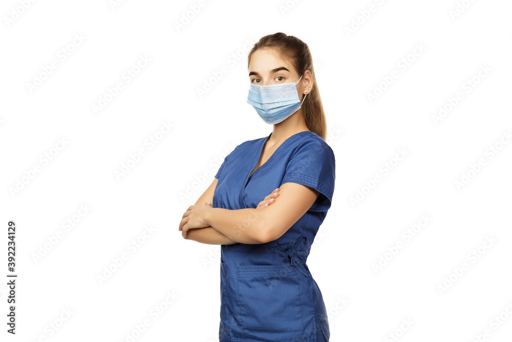 Young beautiful female doctor in blue surgical gown and medical disposable mask on face with folded hands on chest isolated on white Young beautiful female doctor in blue surgical gown and medical disposable mask on face with folded hands on chest isolated on white