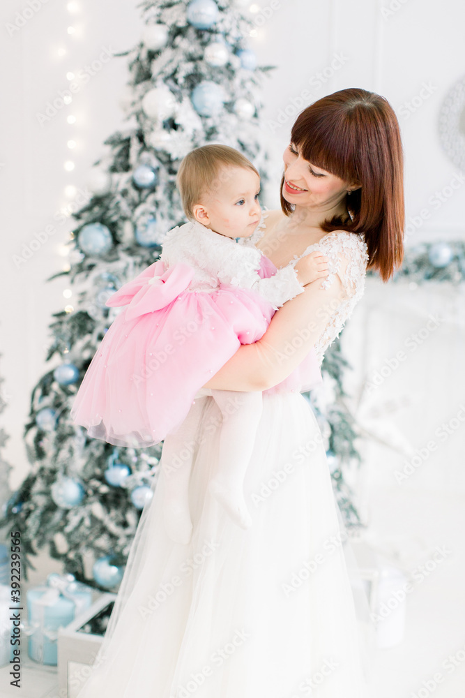 Portrait of smiling young mother holding her beautiful little baby girl, posing on the background of decorated Christmas tree. Family time, winter holidays concept Portrait of smiling young mother holding her beautiful little baby girl, posing on the background of decorated Christmas tree. Family time, winter holidays concept