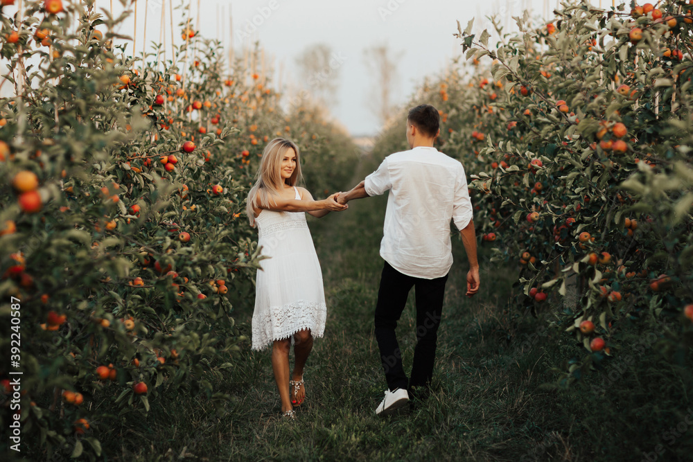 Two lovers in nature. Woman in white dress holding hand of man following her. She is smiling happily.