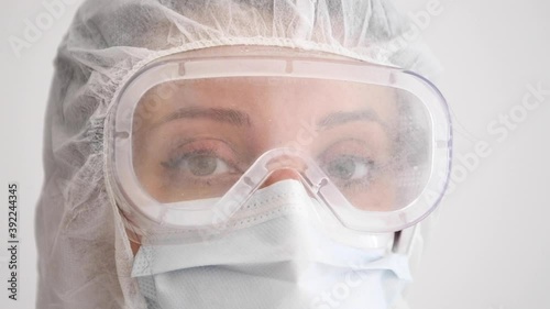 Close up of young female doctor wearing full PPE protective gear against coronavirus covid-19 virus, looks directly into c amera and blinks. Slow motion with white background.