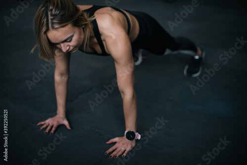 Muscular young woman with perfect athletic body wearing black sportswear doing push-ups exercise on the floor with black mats during sport workout training at modern gym with dark interior.