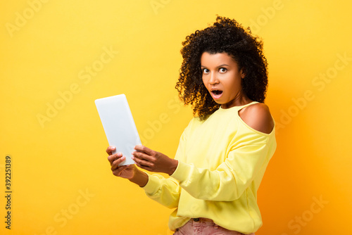 shocked african american woman holding digital tablet and looking at camera on yellow