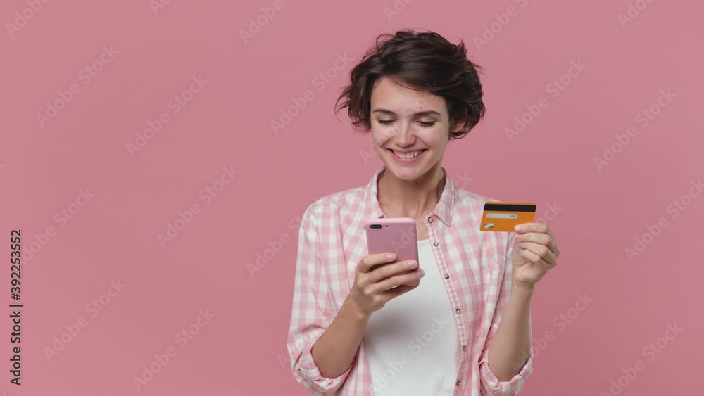 Smiling joyful young woman 20s wearing casual checkered shirt posing isolated on pink background studio. People lifestyle concept. Using mobile cell phone hold credit bank card say yes winner gesture Smiling joyful young woman 20s wearing casual checkered shirt posing isolated on pink background studio. People lifestyle concept. Using mobile cell phone hold credit bank card say yes winner gesture