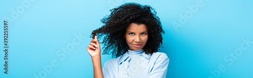 Vászonkép curly african american woman twirling hair on finger on blue, banner