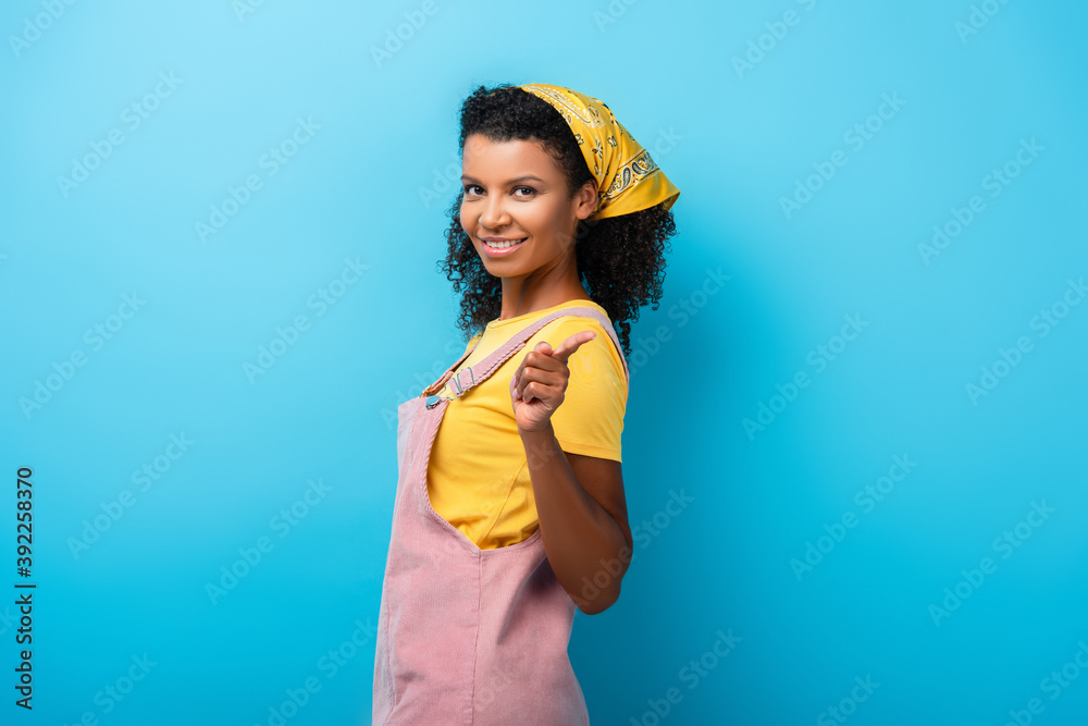 smiling african american woman looking at camera while pointing with finger on blue
