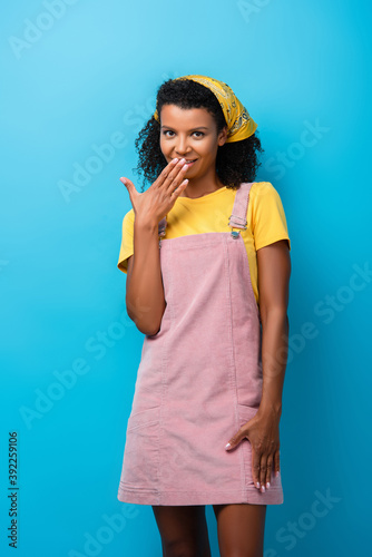 shy african american woman covering mouth with hand isolated on blue