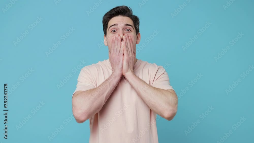 Shocked surprised young man 20s years old in casual beige t-shirt posing isolated on pastel blue colour background in studio. People lifestyle concept. Looking camera say wow covering mouth with hands