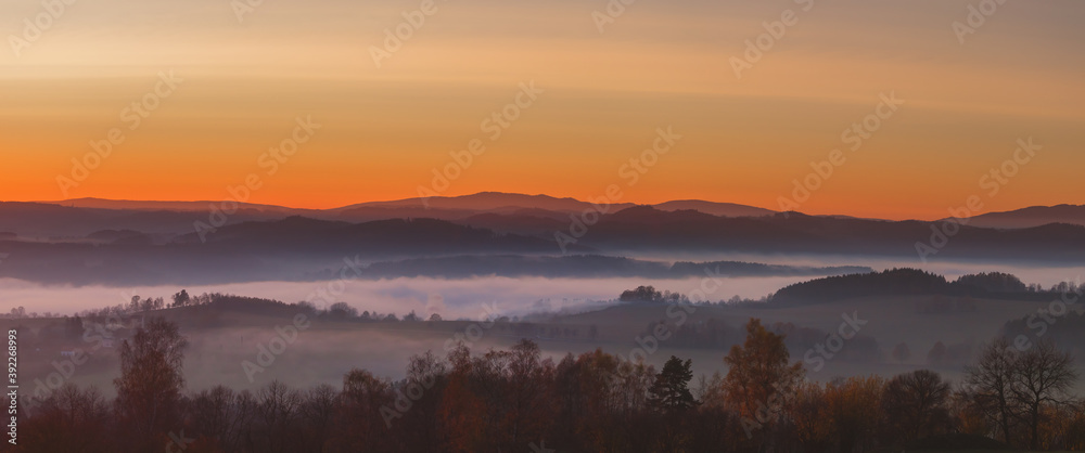 sunset in the mountains - hilly landscape with meadows and forests in a haze, into a yellow and orange-colored sky