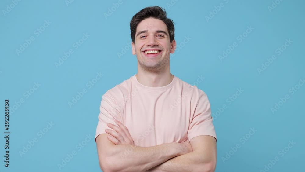 Smiling cheerful handsome young man 20s years old wearing casual beige t-shirt posing isolated on blue background in studio. People lifestyle concept. Looking camera holding hands crossed blinking Smiling cheerful handsome young man 20s years old wearing casual beige t-shirt posing isolated on blue background in studio. People lifestyle concept. Looking camera holding hands crossed blinking