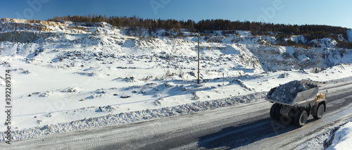 Heavy mining truck transports stone ore against the background of a snow-covered quarry in sunny weather, panorama.