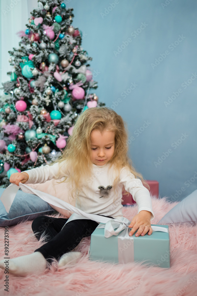 little curly-haired girl sitting on the bed and opening a gift on the background of a Christmas tree little curly-haired girl sitting on the bed and opening a gift on the background of a Christmas tree