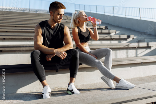 Young sportive couple in sportswear, taking a break to drink water, sitting on the stairs outdoors, healthy lifestyle