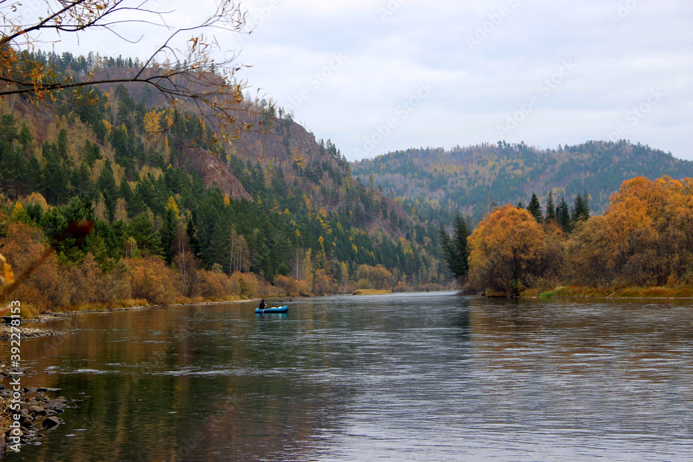 A fast mountain river flows between wooded hills and a lone fisherman on a blue boat. Autumn landscape. A fast mountain river flows between wooded hills and a lone fisherman on a blue boat. Autumn landscape.