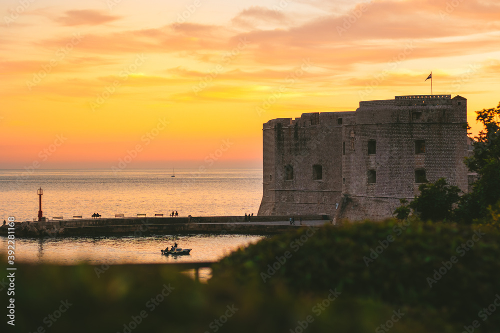 Golden sunset in the town of Dubrovnik, castle standing on the shore of a small harbour, sailboat leaving the harbour and a small local fishing boat entering. Tranquil simple serene setting