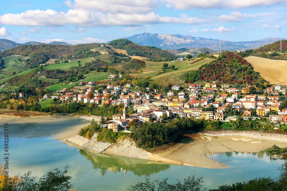 High angel view of small old town on the hill with lake front and surrounded by mountains and colorful autumn trees against nice blue and clouds sky during autumn in Marcatale,The Marche,Italy