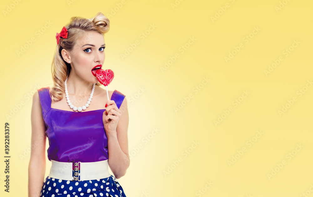Woman eating heart shape lollipop dressed in pinup style dress, with copyspace area for slogan or advertising text message, on yellow background. Pinup girl in retro fashion and vintage studio concept Woman eating heart shape lollipop dressed in pinup style dress, with copyspace area for slogan or advertising text message, on yellow background. Pinup girl in retro fashion and vintage studio concept