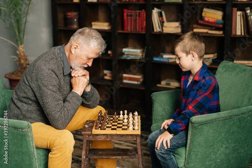 Thoughtful beard grandpa playing chess with grandson, thinking about next step in chess game sitting at chessboard in front of child. Grandchild playing chess with grandparent, leisure activity.