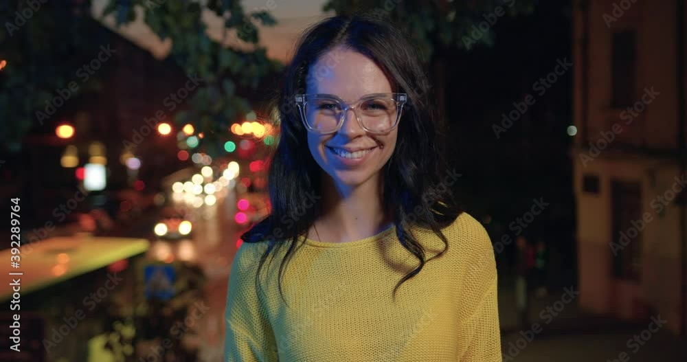 Portrait of charming smiling happy stylish young woman in glasses and casual clothes which poses on camera on lights night city background Portrait of charming smiling happy stylish young woman in glasses and casual clothes which poses on camera on lights night city background