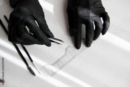 the master in black vinyl gloves holds a pair of tweezers for making eyebrows on the background of a ruler, brushes.