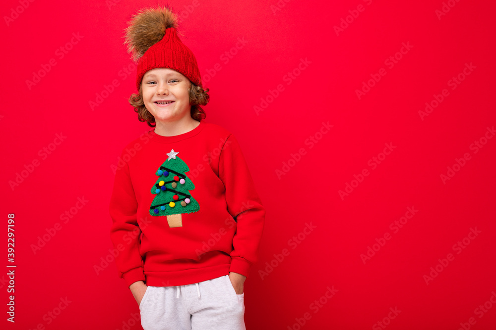 cool blond kid in warm hat and sweater with christmas tree on red background fooling around, christmas concept