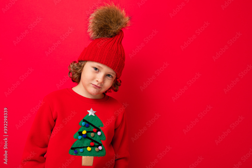 cool blond kid in warm hat and sweater with christmas tree on red background fooling around, christmas concept cool blond kid in warm hat and sweater with christmas tree on red background fooling around, christmas concept