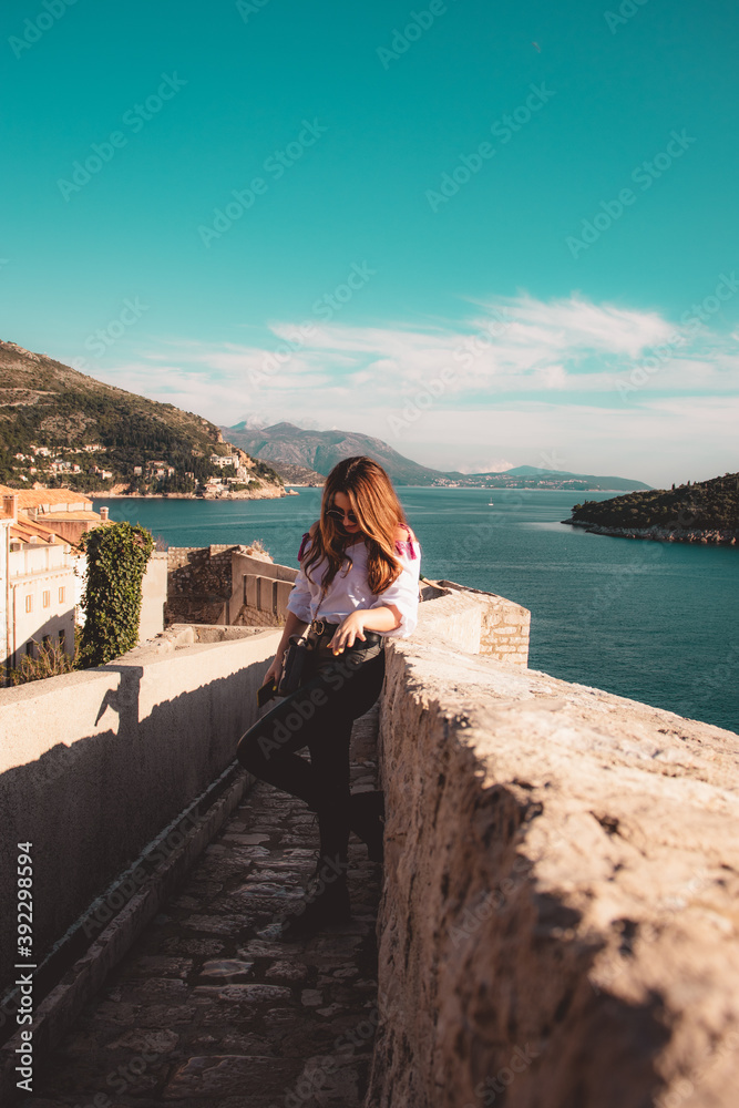 Attractive brunette traveler posing on the old city walls of the town Dubrovnik, tourist exploring the ancient city on a warm autumn day. Wearing a white shirt and black leather pants Attractive brunette traveler posing on the old city walls of the town Dubrovnik, tourist exploring the ancient city on a warm autumn day. Wearing a white shirt and black leather pants