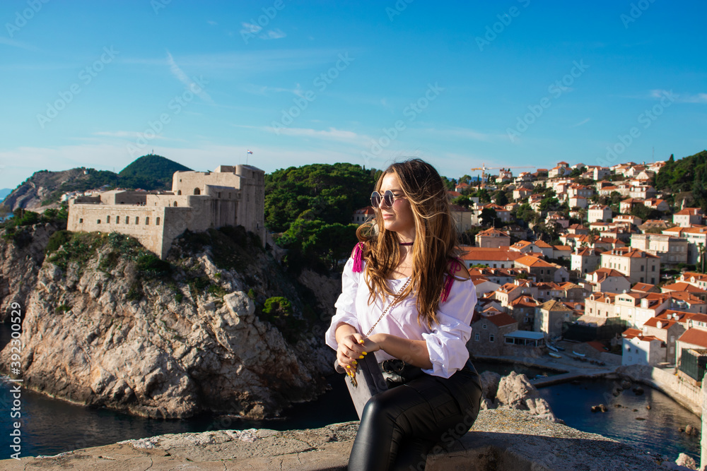 Attractive brunette traveler posing on the old city walls of the town Dubrovnik, tourist exploring the ancient city on a warm autumn day. Wearing a white shirt and black leather pants