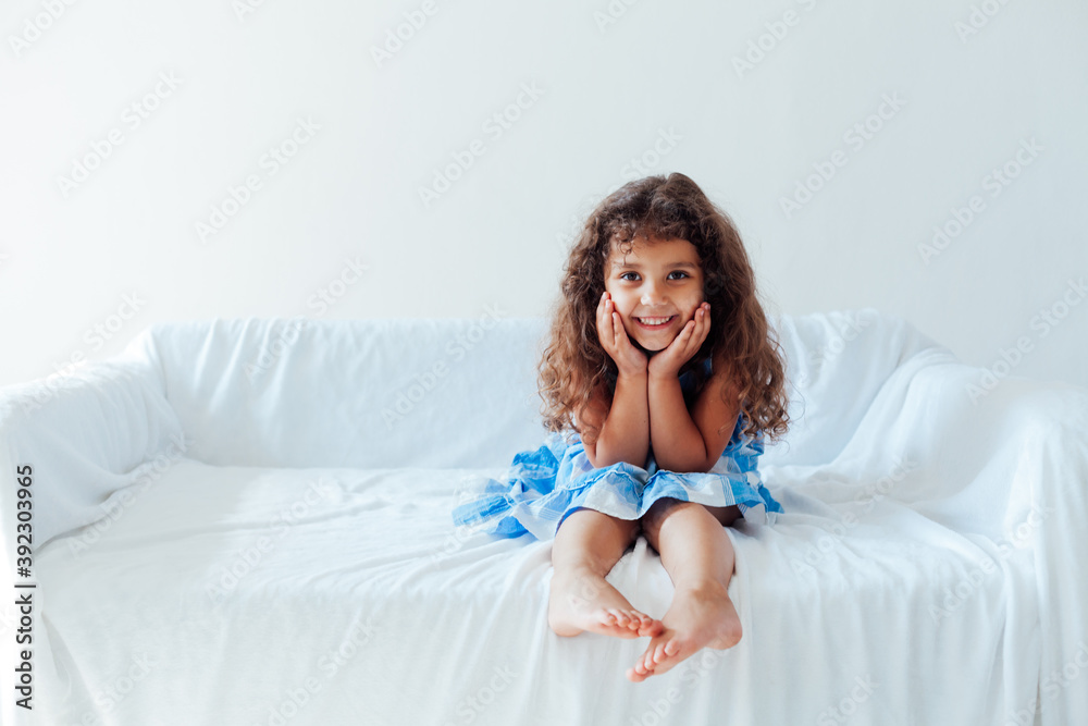 Beautiful little curly girl in a blue dress on the white couch in the room