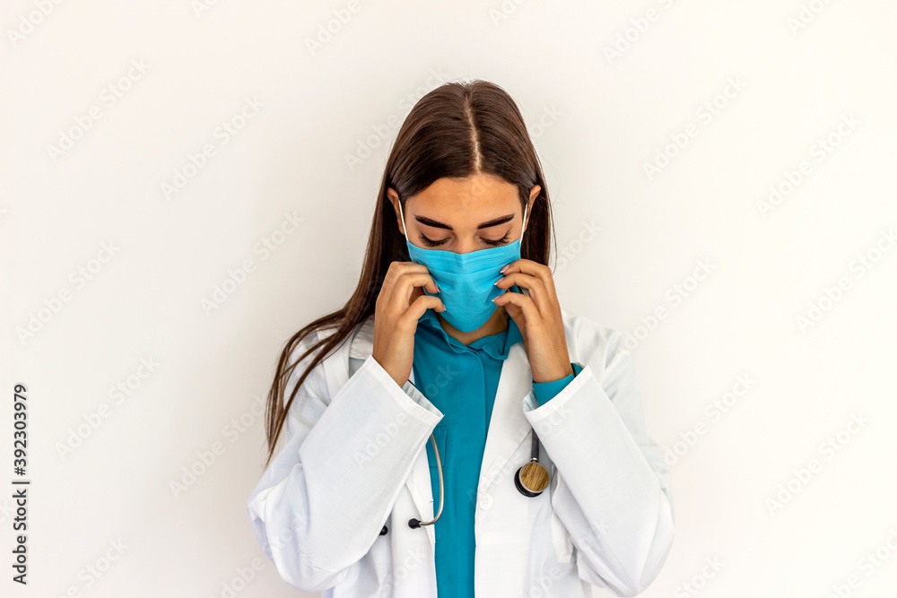 Portrait of a young adult medical professional is wearing lab coat and stethoscope. She is standing against white wall in hospital. Healthcare worker is wearing blue surgical face mask. Copy space. Portrait of a young adult medical professional is wearing lab coat and stethoscope. She is standing against white wall in hospital. Healthcare worker is wearing blue surgical face mask. Copy space.