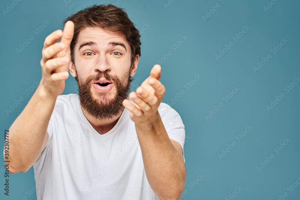 A bearded man in a white T-shirt gestures with his hands emotions blue background A bearded man in a white T-shirt gestures with his hands emotions blue background