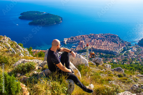 Wide shot from the top of the Srd mountain, man sitting on a rock observing the area around the city of Dubrovnik, orange cablecar descends to the city walls carrying tourist travellers in the summer