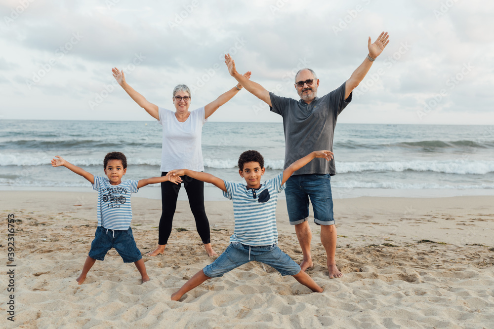 White grandparents with African American grandchildren playing and hugging on the beach with arms raised White grandparents with African American grandchildren playing and hugging on the beach with arms raised