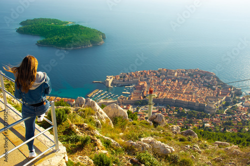 Attractive brunette traveller sitting on a railing above the city of Dubrovnik, viewpoint on Srd mountain. Looking at the ancient historic city surrounded by the blue adriatic sea