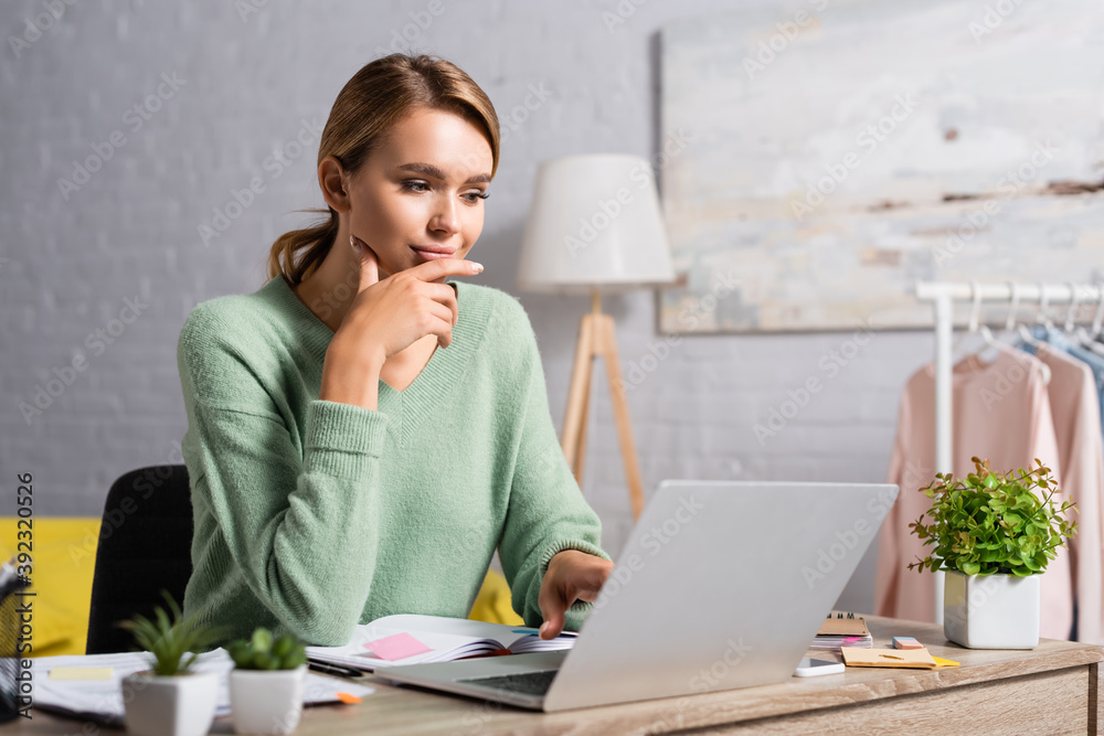 Smiling freelancer using laptop while working near notebook and plants on blurred foreground at home Smiling freelancer using laptop while working near notebook and plants on blurred foreground at home