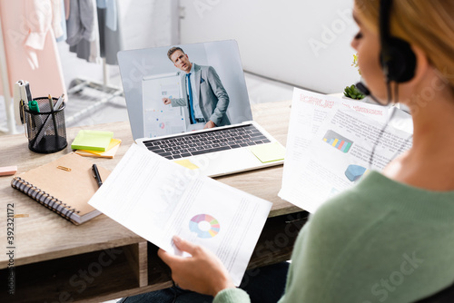 Businessman and flipchart on screen of laptop near freelancer in headset holding papers during video call on blurred foreground