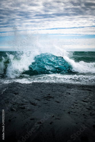 A large chunk of bright blue ice glacier piece on crystal beach in Iceland with waves crashing behind it on a black sand foreground