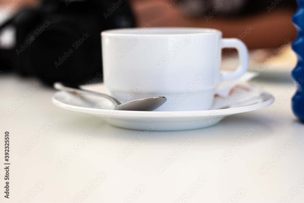 White wide ceramic old fashioned coffee mug standing a white plate with a metal spoon on it White wide ceramic old fashioned coffee mug standing a white plate with a metal spoon on it