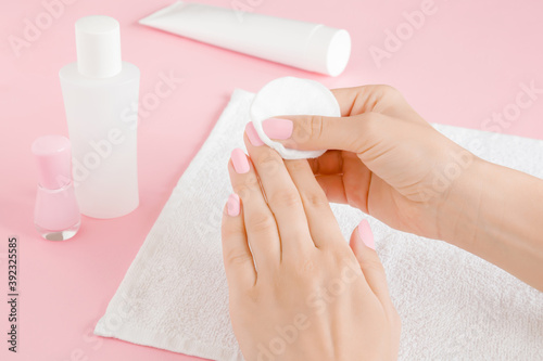 Young adult woman hand removing pink nail polish with white cotton pad on towel on light pink table background. Closeup.