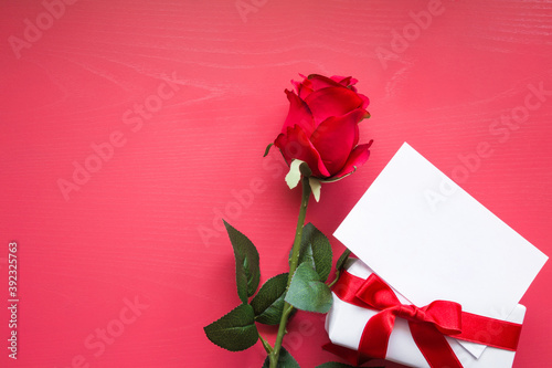 Beautiful red rose, gift box, blank white greeting card on dark pink table background. Lovely, romantic surprise for beloved. Empty place for text, quote or sayings. Top down view. Closeup.
