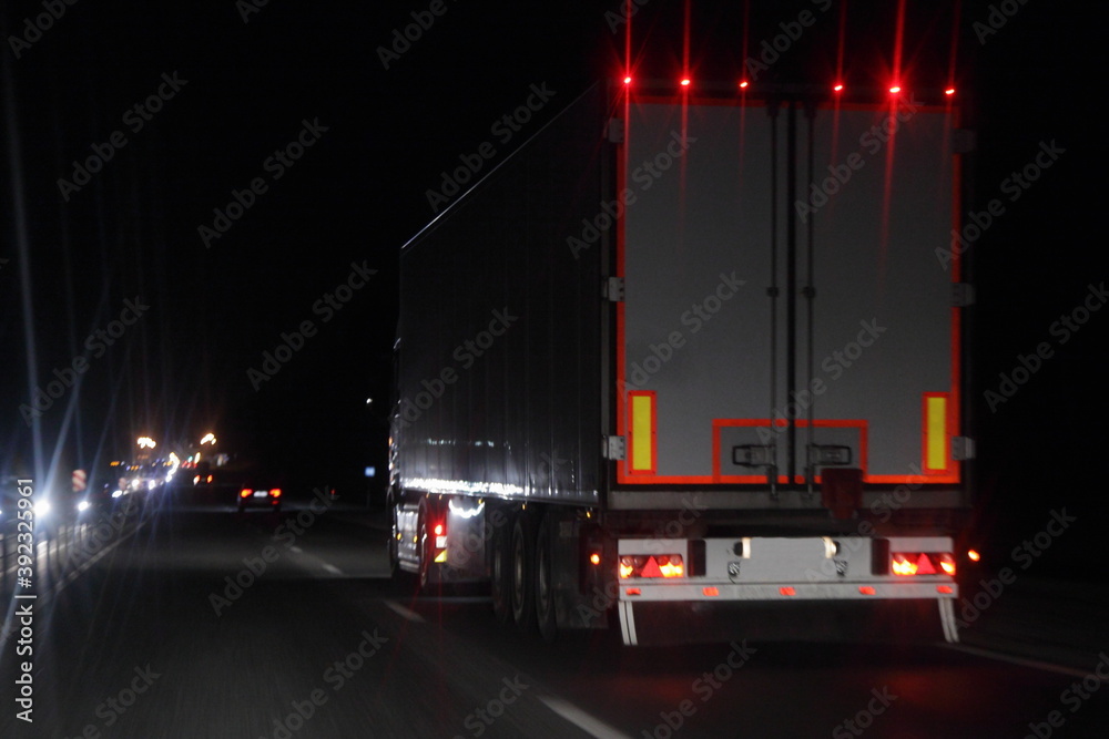 Semi trailer truck with lighting reflection stripe on rear board move on dry asphalted night road in dark, back view - intrenational transportation logistics