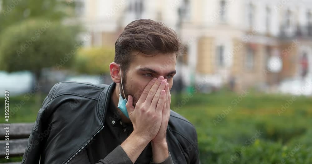 Young sick man in blue medical protective mask sits on a bench in city park, he coughs and blows his nose. Cinema 4K 60fps video Young sick man in blue medical protective mask sits on a bench in city park, he coughs and blows his nose. Cinema 4K 60fps video