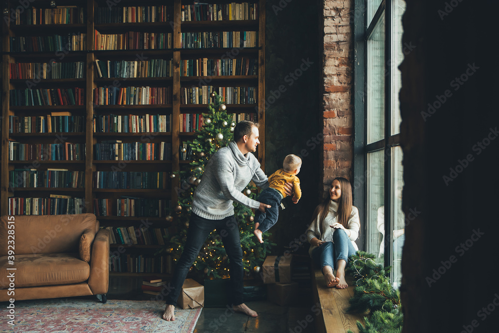 Happy family in cozy living room with decorated Christmas tree. Young father is throwing his little son up, a beautiful mother is laughing