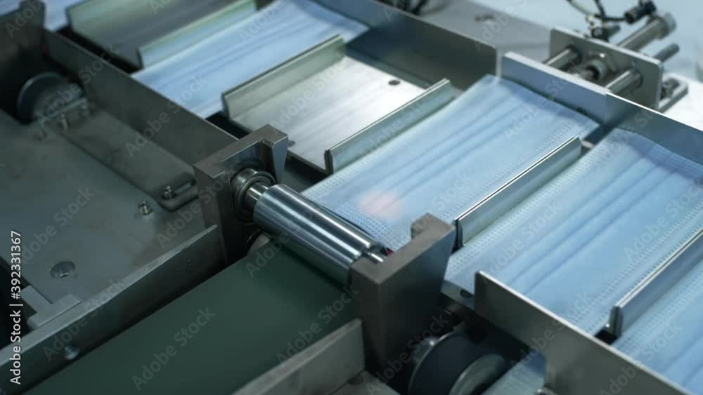Production of medical three-layer blue or white masks. New masks move on a conveyor belt. Face masks for the work of doctors with patients. Making protective masks during the coronavirus pandemic Production of medical three-layer blue or white masks. New masks move on a conveyor belt. Face masks for the work of doctors with patients. Making protective masks during the coronavirus pandemic