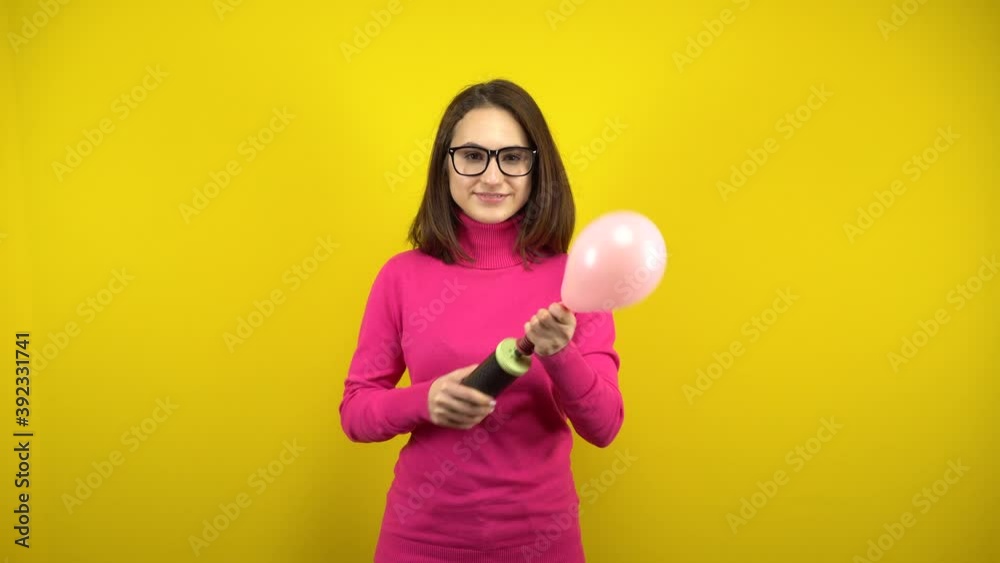A young woman inflates a pink balloon with a pump and it bursts on a yellow background. Girl in a pink turtleneck and glasses. A young woman inflates a pink balloon with a pump and it bursts on a yellow background. Girl in a pink turtleneck and glasses.
