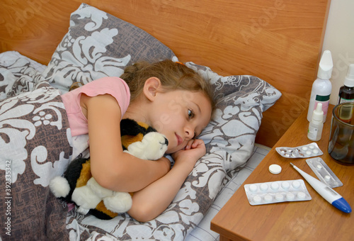 A sad sick girl lies in bed with a soft toy in her hands and looks at the medicine