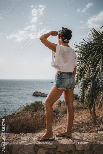 Attractive woman with short hair looking at the sea standing over a cliff and covering the sun from her face with her hand in Ibiza wearing denim shorts