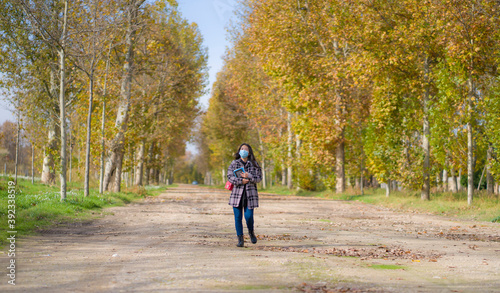 new normal Autumn walk outdoors - young happy and pretty Asian Korean woman in face mask walking cheerful at beautiful city park in vibrant yellow and orange tree leaves © TheVisualsYouNeed