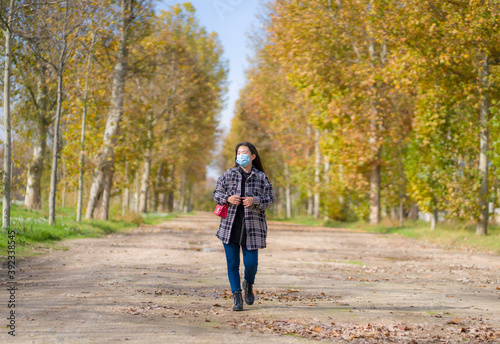 new normal Autumn walk outdoors - young happy and pretty Asian Korean woman in face mask walking cheerful at beautiful city park in vibrant yellow and orange tree leaves