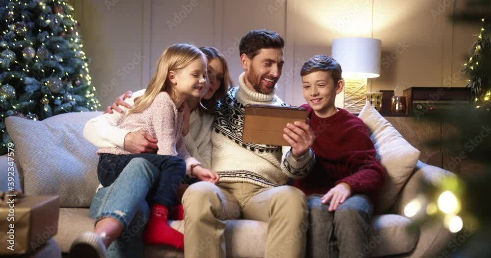 Caucasian cheerful family with children sitting in decorated room with christmas tree and videochatting on tablet Parents with kids speaking on video call on New Year's Eve. Holidays concept Caucasian cheerful family with children sitting in decorated room with christmas tree and videochatting on tablet Parents with kids speaking on video call on New Year's Eve. Holidays concept