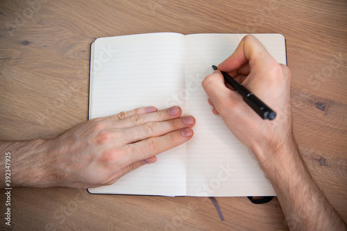 Top view of male hands with a pen on a notebook on a wooden desk.